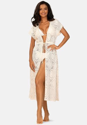 Kobieta w białym bikini i przezroczystej, wzorzystej, sięgającej do kolan narzutce plażowej, uśmiechająca się z jedną ręką na biodrze.