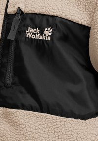 Μπεζ φλις μπουφάν με μαύρη τσέπη στο στήθος με φερμουάρ που φέρει το λογότυπο Jack Wolfskin και αποτύπωση πατούρας σε λευκή κέντηση.