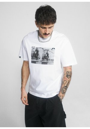 Mężczyzna z tatuażami w białym koszulce z nadrukiem i czarnych spodniach, patrzący w dół na jednolite jasne tło.