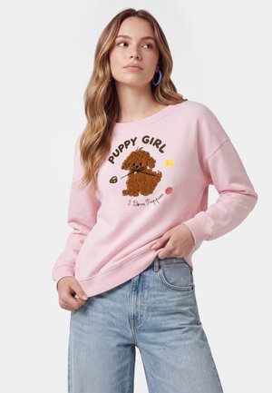 Kvinde med langt bølget hår iført blå hoop-øreringe, lyserød sweatshirt med brun hvalp og tekst "Puppy Girl I Love Puppies," samt blå jeans.