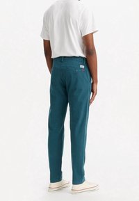 Turkusowe bawełniane spodnie o prostych nogawkach, z tylną kieszenią, zapięciem na guzik oraz białą metką. Noszone z białym t-shirtem.