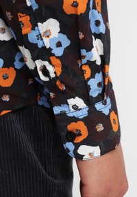 Camisa floral con fondo negro y flores naranjas, azules y blancas, con puños abotonados y detalles de tela texturizada.