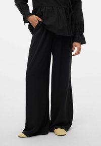 Luźne czarne spodnie o szerokich nogawkach wykonane z teksturowanej tkaniny, zestawione z luźną czarną bluzką i żółtymi butami wsuwanymi. Charakterystyczne detale to swobodny krój.