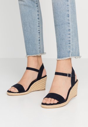 Kvinde iført lyseblå jeans med frynset kant og sorte kile-sandaler med ankelrem og vævet platformsål, stående på en hvid gulv.