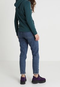 Felpa verde scuro con cappuccio, jeans blu con orli risvoltati e sneakers in suede viola con suole nere spesse, viste da dietro.