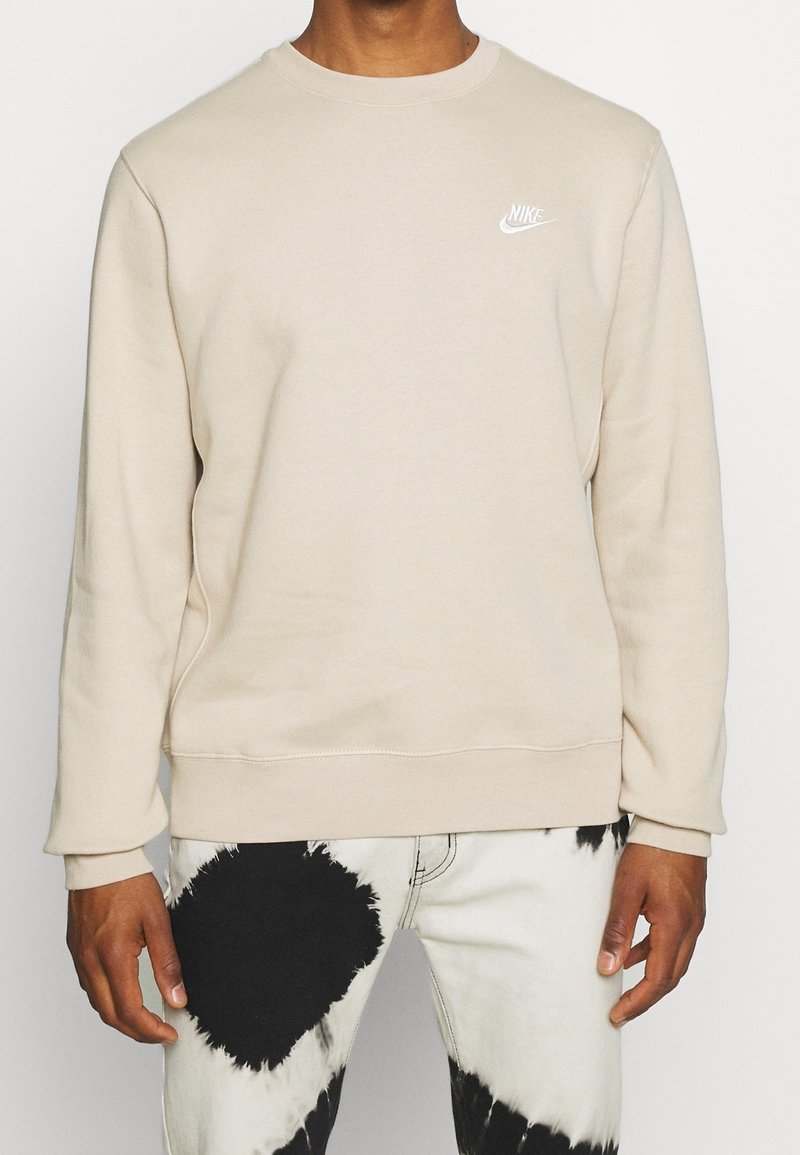 Personne portant un sweat-shirt Nike beige et un jean blanc avec de grandes taches noires irrégulières, debout devant un fond uni.