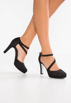 Nohy v černých třpytivých lodičkách na vysokém podpatku typu Mary Jane s pásky kolem kotníku a střední části chodidla, stojící na bílém podlaze.