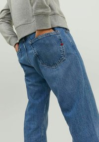 Hellblaue Jeans aus Denim mit einem verwaschenen Finish, ausgestattet mit zwei Gesäßtaschen und einem kleinen roten Akzent auf der rechten Tasche. Kombiniert mit einem grauen Sweatshirt.