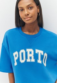 Zils džemperis ar vaļēju piegriezumu un priekšpusē baltu uzrakstu "PORTO". Izgatavots no mīkstas auduma ar gludu tekstūru un ribota izgriezuma kaklu.