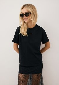 Czarna bawełniana koszulka z krótkim rękawem i okrągłym dekoltem, w połączeniu z czarną koronkową spódnicą. Dodatki to okrągłe okulary przeciwsłoneczne.