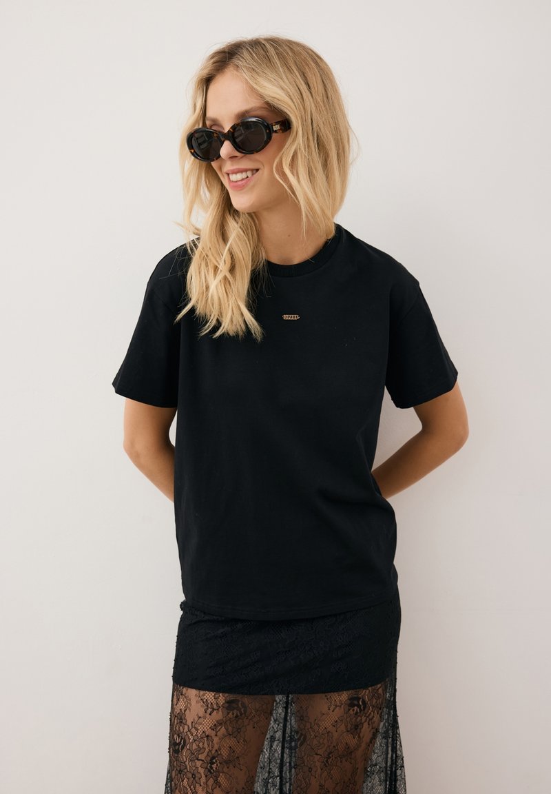 Czarna bawełniana koszulka z krótkim rękawem i okrągłym dekoltem, w połączeniu z czarną koronkową spódnicą. Dodatki to okrągłe okulary przeciwsłoneczne.