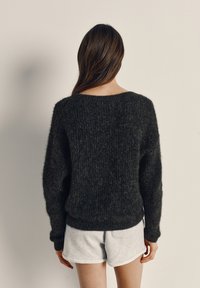 Sieviete ar gariem brūniem matiem, valkājot tumšu pūkainu džemperi un gaiši pelēkus šortus, stāvot pret vienkāršu gaiši krēmkrāsas sienu.