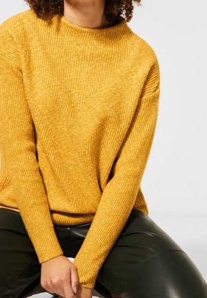Personne portant un pull côtelé jaune moutarde et un pantalon en cuir noir, assise les mains jointes devant sur un fond clair.