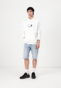 Sweatshirt à capuche blanche avec un graphique à l'avant, associé à un short en denim bleu clair et des baskets noires avec des accents blancs et des chaussettes.