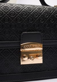 Sac à main en cuir noir avec un motif en diamants, fermoir en or avec la marque "ANNA FIELD" et des accents décoratifs circulaires.