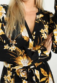 Robe noire avec un motif floral jaune et blanc. Présente un devant à boutons, des manches longues et un détail de taille nouée.