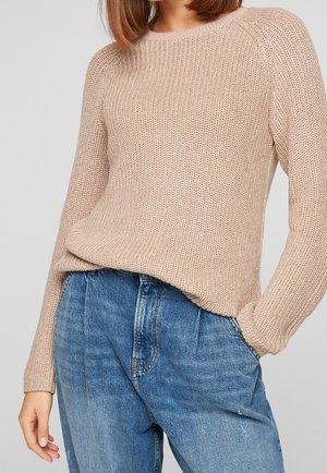 Femme portant un pull en tricot beige et un jean en denim bleu taille haute, avec une main dans la poche, sur un fond uni.