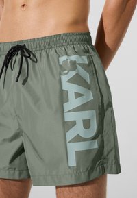 Zöld úszónadrág fekete zsinórral a deréknál, nagy függőleges világoskék "KARL" logóval a bal combon, amit egy személy visel.