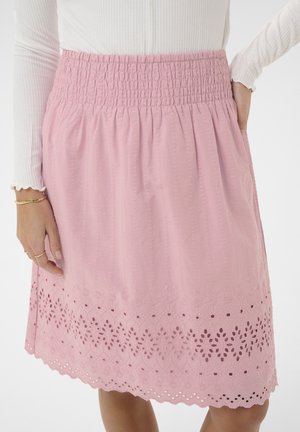 Mujer con falda rosa con cintura fruncida y bordado floral calado cerca del dobladillo en forma de ondas, combinada con una camiseta blanca de manga larga con tejido acanalado.