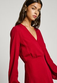 Robe portefeuille rouge à manches longues, avec un tissu texturé et un décolleté en V. La taille est froncée, mettant en valeur la silhouette.