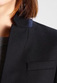 Blazer negro a medida con cuello estructurado y un acento en azul marino en el escote. Tejido texturizado con una silueta ajustada y entallada.
