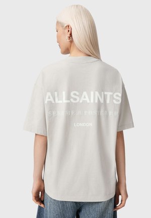 Vrouw met lang blond haar die een licht beige oversized AllSaints T-shirt draagt met bedrukt tekst op de achterkant en een blauwe spijkerbroek.
