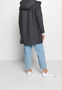 Lång svart vattentät jacka med huva, med en mjuk textur, i kombination med ljusblå denimbyxor och vita sneakers.