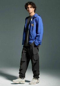 Giacca leggera blu con zip frontale, dettaglio grafico, t-shirt nera, pantaloni cargo neri e sneakers beige testurizzate.