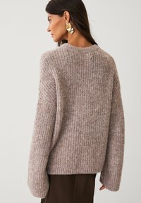 Overdimensioneret strikket sweater i blød taupe med ribbet tekstur, brede ærmer og en rund halsudskæring. Båret over mørkebrune bukser.