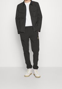 Černá bunda se dvěma předními kapsami, spárovaná s černými kalhotami na stažení se znakem. Pod tím bílá tričko a bílé tenisky.