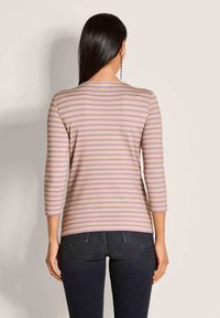 Vrouw met lang zwart haar die een beige en roze gestreepte lange mouwen top en donker skinny jeans draagt, met haar gezicht naar een effen muur.