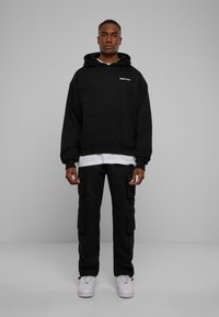 Felpa nera con cappuccio e tasca frontale, abbinata a pantaloni cargo neri con tasche laterali. Sneakers bianche completano il look.