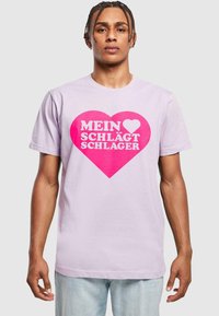 Heledast lilla puuvillane T-särk, millel on suur roosa südame graafika, millel on valge tekst "MEIN ♥ SCHLÄGT SCHLAGER." Lühikesed varrukad ja klassikaline lõige.