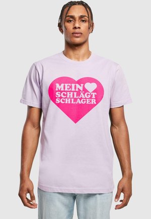 Lys lilla bomulds t-shirt med et stort pink hjertegrafik med hvid tekst, der står "MEIN ♥ SCHLÄGT SCHLAGER." Korte ærmer og klassisk pasform.