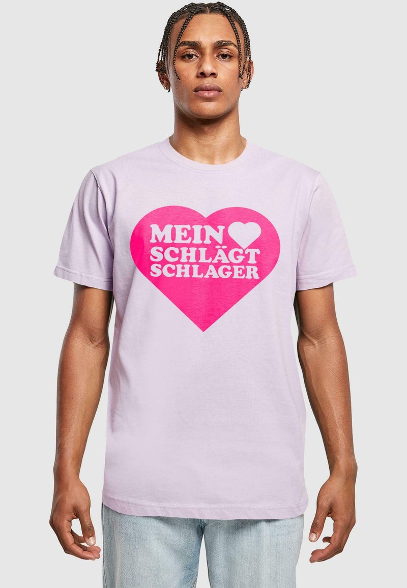 Heledast lilla puuvillane T-särk, millel on suur roosa südame graafika, millel on valge tekst "MEIN ♥ SCHLÄGT SCHLAGER." Lühikesed varrukad ja klassikaline lõige.