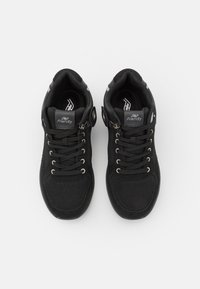 Des chaussures de sport noires avec une tige en maille, des lacets ronds et des œillets métalliques. Elles présentent un logo sur la languette et des accents réfléchissants sur les côtés.
