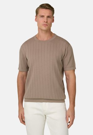 Man draagt een beige geribbelde korte mouw trui en witte broek, staand tegen een effen lichte achtergrond.