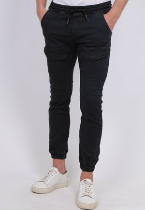 Homme portant un pantalon de jogging cargo noir avec des poignets élastiques et des baskets blanches, les mains dans les poches, debout devant un fond uni.