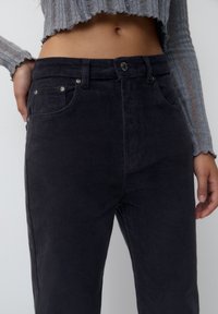 Pantalon en velours côtelé noir avec une coupe droite, comportant des poches avant et des garnitures en métal argenté à la taille et aux poches. Surface texturée.
