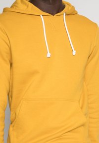 Hombre con una sudadera color mostaza con cordones blancos y bolsillo tipo canguro en la parte frontal, sobre un fondo claro.