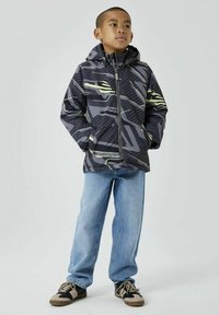 Giacca nera imbottita con motivi geometrici grigi e gialli, con zip frontale e cappuccio; abbinata a jeans azzurri chiari e scarpe nere.