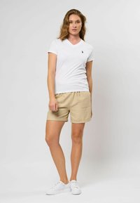 Witte V-hals t-shirt, beige shorts met elastische tailleband, opgerolde zoom en twee achterzakken. Model draagt witte sneakers.