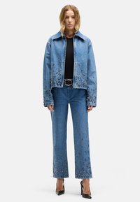 Kurtka dżinsowa i szerokie spodnie dżinsowe, oba ozdobione metalowymi ćwiekami. Kurtka ma rozłożysty kołnierz, jest jasnoniebieska i ma prosty krój.