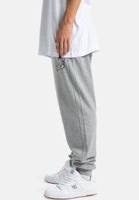 Graue Fleece-Jogginghose mit einem Logo auf dem Oberschenkel, kombiniert mit weißen Sneakers. Das Shirt ist schlicht weiß, und die Hände sind in den Taschen.