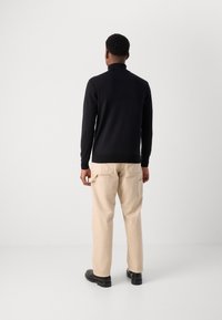 Pull-over noir à manches longues avec col montant, associé à un pantalon cargo beige et des chaussures noires. Texture lisse, coupe ajustée.