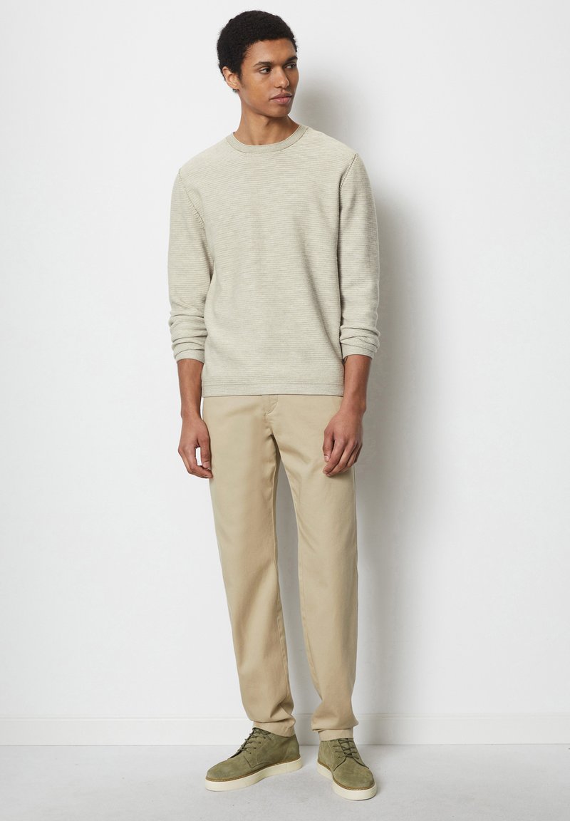 Jasnoszara teksturowana bluza z ribbingu, zestawiona z beżowymi spodniami i zielonymi zamszowymi butami. Model stoi na jednolitym tle.