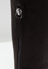 Botte en daim noir avec une fermeture éclair latérale à tirette argentée, texture lisse et design ajusté sur la partie supérieure.