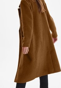 Brun uldfrakke med en struktureret overflade, lange ærmer, afslappet pasform og sidelæg. Frakken når under knæet og har et enkelt design.