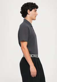 Mørkegrå polo skjorte med klassisk krage, korte ærmer, parret med sorte bukser med en kontrasterende hvid logo elastik.