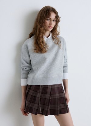 Junge Frau mit langen lockigen Haaren, die einen grauen Sweatshirt über einem weißen Hemd und einen plissierten braunen karierten Rock mit Kettendetail trägt.